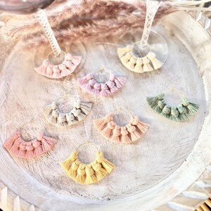 May include: A collection of handmade macrame wine glass charms in various pastel colors, including pink, yellow, and green. Each charm features a gold-colored hoop and a fan-shaped design, perfect for adding a decorative touch to glassware.