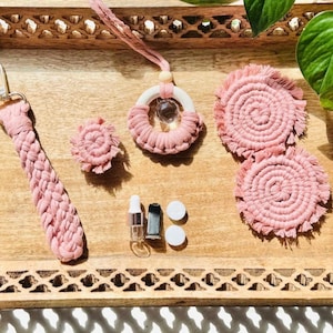 May include: A collection of handmade macrame accessories in shades of pink, including a keychain, a car air freshener, a small flower, two coasters, and two small bottles. The items are arranged on a wooden tray.