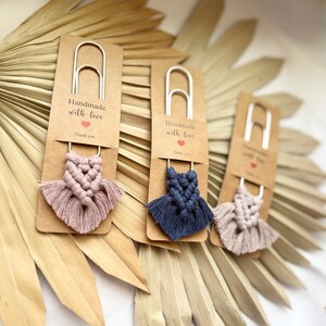 May include: Three handmade macrame bookmarks in shades of pink, blue, and beige. Each bookmark features a silver paperclip and a small macrame design. The cards read "Handmade with love" and "Thank you."