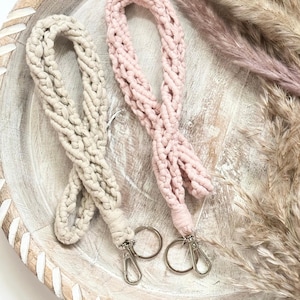 May include: Two macrame lanyards in the image. One is a light beige color, and the other is a soft pink. Each lanyard has a silver-colored clasp and a key ring. The lanyards are displayed on a wooden tray with dried decorative plants.