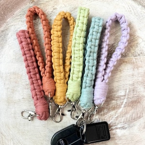 May include: A collection of six macrame wrist lanyards in a variety of colors, including pink, orange, yellow, green, blue, and purple. Each lanyard is attached to a silver-colored clasp. A black car key fob is attached to one of the lanyards.