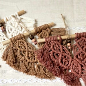 May include: Close-up of several macrame wall hangings in shades of brown and cream. Each piece features intricate knotting, a wooden dowel, and decorative beads. The fringe adds texture and visual interest to these handcrafted home decor items.