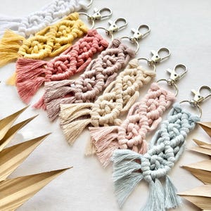 May include: A set of seven colorful macrame keychains with silver key rings. The keychains are made with a variety of colors, including pink, blue, yellow, and white. Each keychain has a tassel at the bottom.