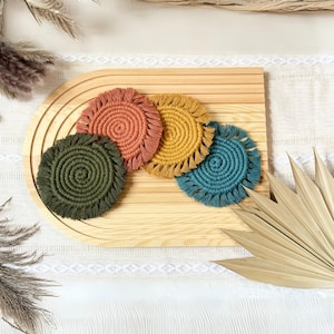 May include: Four round crocheted coasters with fringed edges in shades of teal, mustard yellow, and burnt orange. The coasters are arranged on a wooden surface.