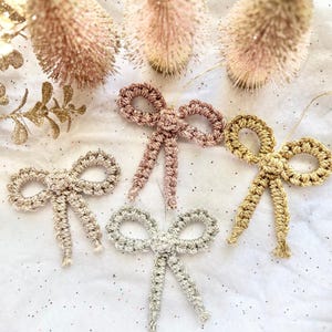 May include: Four handmade crochet bow ornaments in shades of gold, silver, and rose. The bows are arranged on a white surface with glitter and decorative gold and pink accents. These ornaments are perfect for holiday decor.
