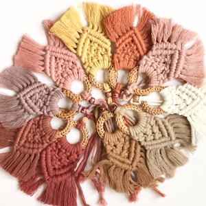 May include: A collection of handmade macrame coasters in various muted colors, including pink, beige, and yellow. Each coaster features a woven design with a fringed edge and a natural wooden ring. The coasters are arranged in a circular pattern.