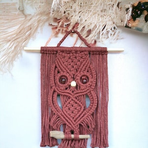 May include: A handmade macrame owl wall hanging in a muted reddish-brown color. The owl features wooden bead eyes and a small wooden beak. The macrame design includes intricate knotting and fringe, mounted on a natural wooden dowel for easy hanging.