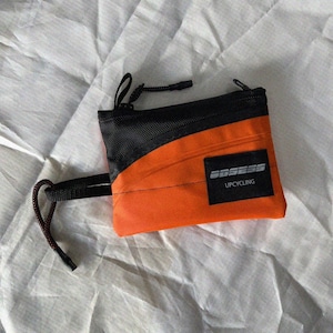 Könnte beinhalten: Eine rechteckige Reißverschlusstasche mit einem leuchtend orangefarbenen unteren Bereich und einem schwarzen Netzeinsatz oben. Ein schwarzer rechteckiger Aufnäher auf der Vorderseite zeigt den weißen Text "OBSESS" über dem Wort "UPCYCLING". Eine schwarze Kordelschlaufe ist an der Seite befestigt.