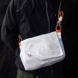 May include: White crossbody bag with orange detailing and a matching orange rope strap. The bag features a flap closure and the text "ASSESS UPCYCLING". The bag is made of a lightweight material.