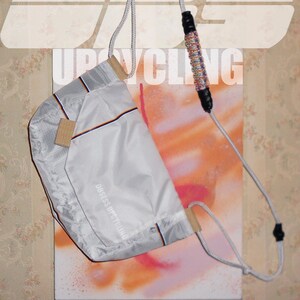 May include: A light grey upcycled bag with a drawstring closure and a shoulder strap. The bag has a rectangular shape with a beige accent and a thin orange and black stripe. The words "OBSESS UPCYCLING" are printed on the side. The background has the word "UPCYCLING" and the date "02/26".