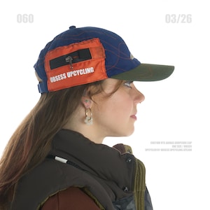 Op de afbeelding: Een marineblauwe en oranje baseballcap met de tekst "OBSESS UPCYCLING". De cap heeft een groene klep en een klein oranje zakje met een rits. One size en unisex.