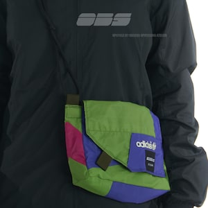 Può includere: Una borsa a tracolla multicolore con pannelli verdi, viola e rosa. La borsa presenta il logo Adidas su una patta viola e un cinturino nero. La persona indossa una giacca nera. I testi "03/26" e "OBS" sono visibili.