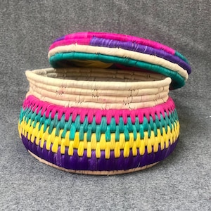 Può includere: Un cestino rotondo intrecciato con coperchio, caratterizzato da vivaci fasce colorate. Il corpo del cestino presenta tonalità rosa, verdi, gialle e viola, mentre il coperchio rispecchia questi colori. Ideale per l'arredamento.