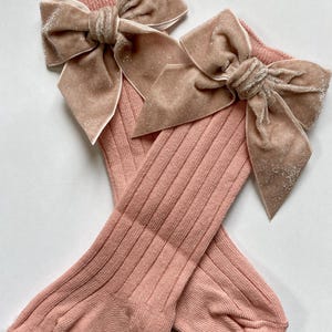 May include: A pair of pink knee-high socks with a ribbed texture. Each sock features a large, sparkly, velvet bow in a complementary shade of beige. The bows are centered at the top of the socks, adding a decorative touch.