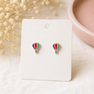 May include: A pair of stud earrings featuring a hot air balloon design. The balloons have a rainbow color scheme and are set against a silver-tone metal. The earrings are displayed on a white card.
