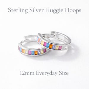 May include: Sterling silver huggie hoops with colorful rectangular gemstones. The earrings are 12mm in size. The gemstones are arranged in a rainbow pattern, with colors including pink, orange, yellow, blue, and purple. The earrings are on a white background.