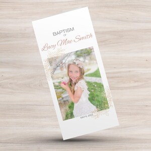 Peut inclure: Une carte blanche avec des motifs floraux dorés et le texte "Bapteme de Lucy Mae Smith" en rose. La carte comprend également une photo d'une jeune fille en robe blanche tenant une fleur, avec la date "May 6, 2023" imprimée sous la photo.