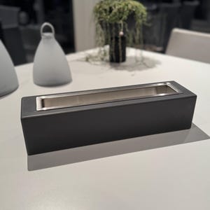 Puede incluir: Una jardinera rectangular negra mate con interior de acero inoxidable. La jardinera est&aacute; sobre una superficie blanca, con un fondo borroso con objetos decorativos y vegetaci&oacute;n. El dise&ntilde;o es moderno y minimalista.