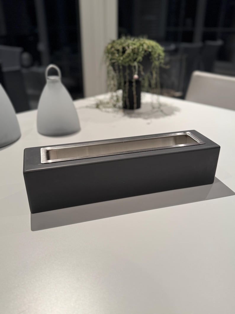 Puede incluir: Una jardinera rectangular negra mate con interior de acero inoxidable. La jardinera est&aacute; sobre una superficie blanca, con un fondo borroso con objetos decorativos y vegetaci&oacute;n. El dise&ntilde;o es moderno y minimalista.