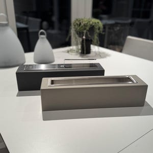Puede incluir: Dos jardineras rectangulares de estilo moderno. Una es negra, la otra es de color topo claro. Ambas tienen un dep&oacute;sito interior de acero inoxidable. Las jardineras est&aacute;n sobre una superficie blanca, con otros objetos decorativos en el fondo.