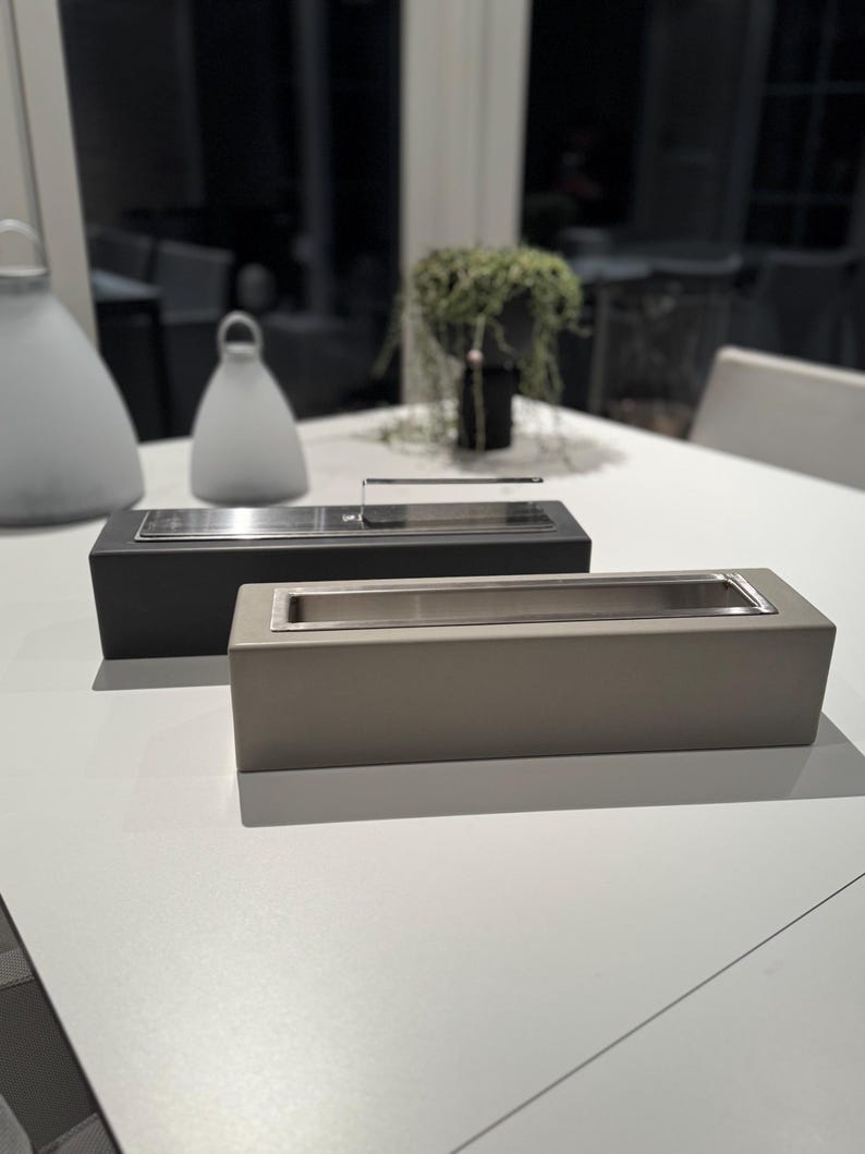 Puede incluir: Dos jardineras rectangulares de estilo moderno. Una es negra, la otra es de color topo claro. Ambas tienen un dep&oacute;sito interior de acero inoxidable. Las jardineras est&aacute;n sobre una superficie blanca, con otros objetos decorativos en el fondo.