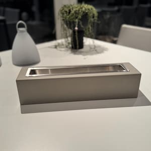 Puede incluir: Un brasero rectangular gris claro con interior de acero inoxidable. El brasero est&aacute; sobre una superficie blanca. Un objeto blanco en forma de campana y una jardinera negra con vegetaci&oacute;n est&aacute;n en el fondo.