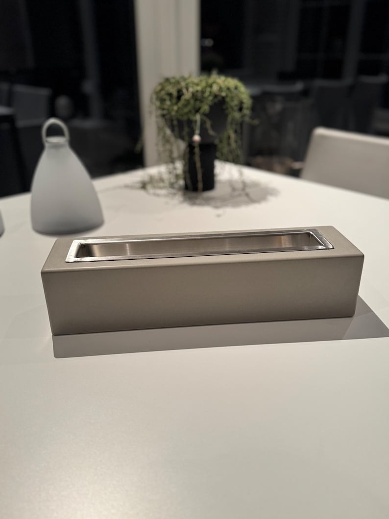 Puede incluir: Un brasero rectangular gris claro con interior de acero inoxidable. El brasero est&aacute; sobre una superficie blanca. Un objeto blanco en forma de campana y una jardinera negra con vegetaci&oacute;n est&aacute;n en el fondo.