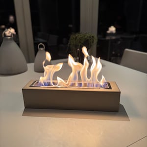 Puede incluir: Una chimenea de mesa rectangular beige con llamas danzantes. Las llamas son amarillas y naranjas con azul en la base. La chimenea est&aacute; sobre una mesa blanca, con otros objetos decorativos en el fondo.