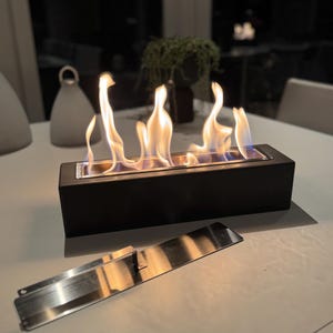 Puede incluir: Una chimenea de mesa rectangular negra con llamas. Una placa met&aacute;lica de acero inoxidable est&aacute; delante de la chimenea. Las llamas son amarillas y naranjas. La chimenea est&aacute; sobre una mesa blanca.