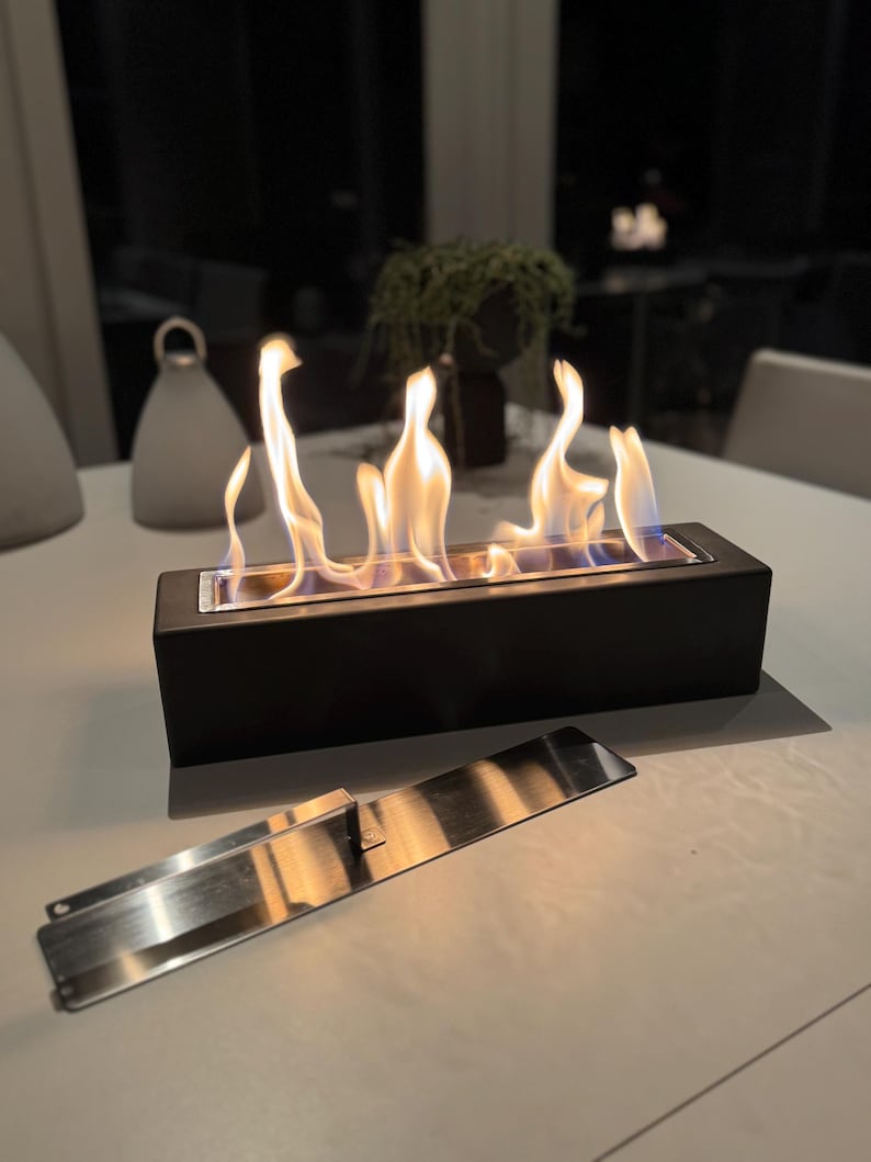 Puede incluir: Una chimenea de mesa rectangular negra con llamas. Una placa met&aacute;lica de acero inoxidable est&aacute; delante de la chimenea. Las llamas son amarillas y naranjas. La chimenea est&aacute; sobre una mesa blanca.