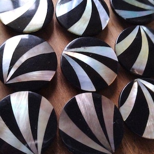 May include: A collection of round, black buttons with a radial design of alternating black and iridescent white stripes. The buttons are arranged closely together, showcasing their decorative pattern and potential for craft projects.