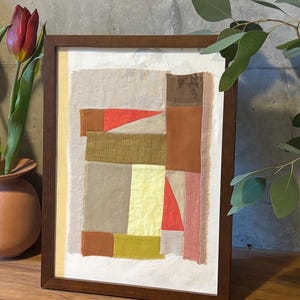 以下が含まれることがあります： 茶色、ベージュ、ピンクの布を様々な色合いで組み合わせたパッチワークのデザインが施された、額装された抽象的な芸術作品。布は幾何学的なパターンで配置されています。