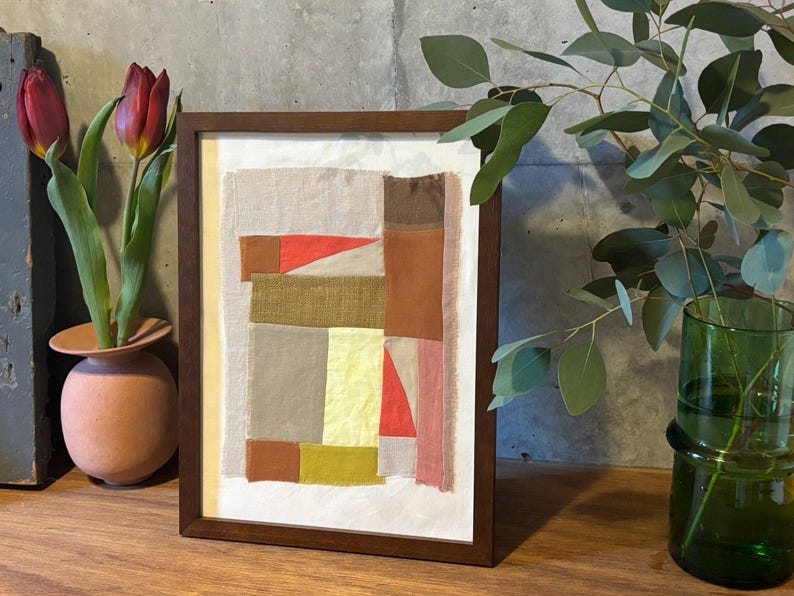 May include: A framed abstract art piece with a patchwork design of various colors and textures, including brown, red, yellow, and white. The frame is dark brown wood.