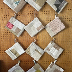 May include: Twelve small fabric pouches with different patchwork designs, hanging on a pegboard wall. The pouches are made of beige linen fabric with various patterns in beige, brown, red, and white.