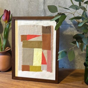 May include: A framed abstract art piece with a patchwork design of various colors and textures, including brown, red, yellow, and white. The artwork is displayed on a wooden surface.