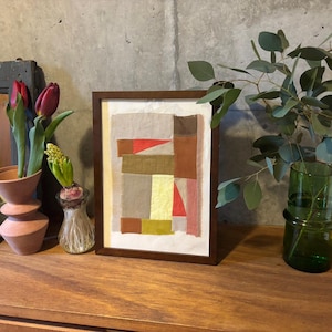 May include: A framed abstract art print with a geometric design in shades of brown, red, and yellow. The print is displayed on a wooden shelf with a vase of flowers and a potted plant.