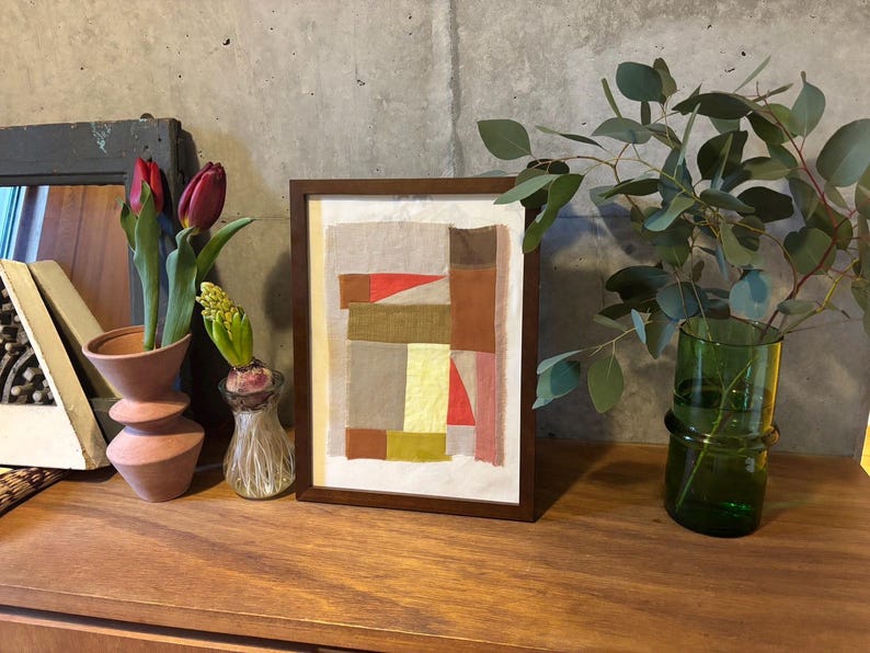 May include: A framed abstract art print with a geometric design in shades of brown, red, and yellow. The print is displayed on a wooden shelf with a vase of flowers and a potted plant.