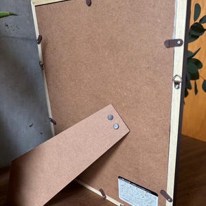 May include: The back of a brown wooden picture frame with a brown cardboard backing. The frame has a built-in easel stand and a small label with Japanese text.