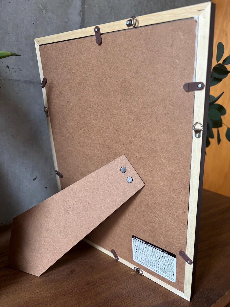 May include: The back of a brown wooden picture frame with a brown cardboard backing. The frame has a built-in easel stand and a small label with Japanese text.