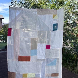 Könnte beinhalten: Ein rechteckiger Wandbehang oder Quilt aus Textil mit einem Patchwork-Design aus halbtransparenten weißen Stoffbahnen, akzentuiert durch verschiedene einfarbige rechteckige und dreieckige Flicken in den Farben Rosa, Blau, Braun, Gelb und Grau.