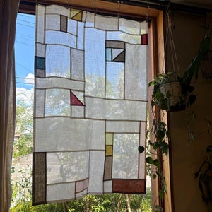 Puede incluir: Una cortina de lino blanco con un patrón geométrico de cuadrados y rectángulos coloridos. La cortina está colgada en una ventana con vista a una terraza de madera y vegetación verde.