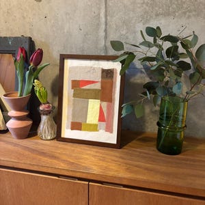 May include: A framed abstract art print with a collage of colorful paper squares and rectangles in shades of brown, red, yellow, and beige. The print is displayed on a wooden shelf with a vase of flowers and a potted plant.