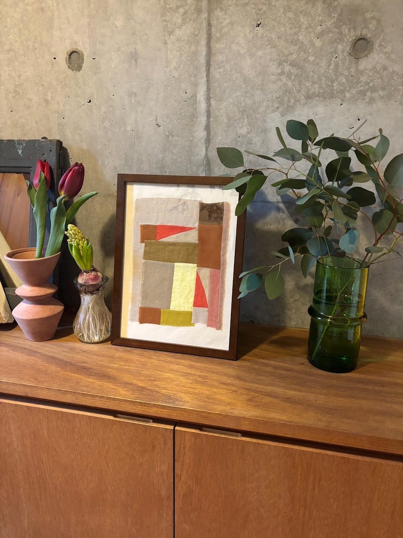 May include: A framed abstract art print with a collage of colorful paper squares and rectangles in shades of brown, red, yellow, and beige. The print is displayed on a wooden shelf with a vase of flowers and a potted plant.