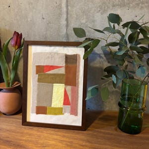May include: A framed abstract art piece with a patchwork design of various shades of brown, beige, yellow, pink, and red fabric. The frame is dark brown wood.