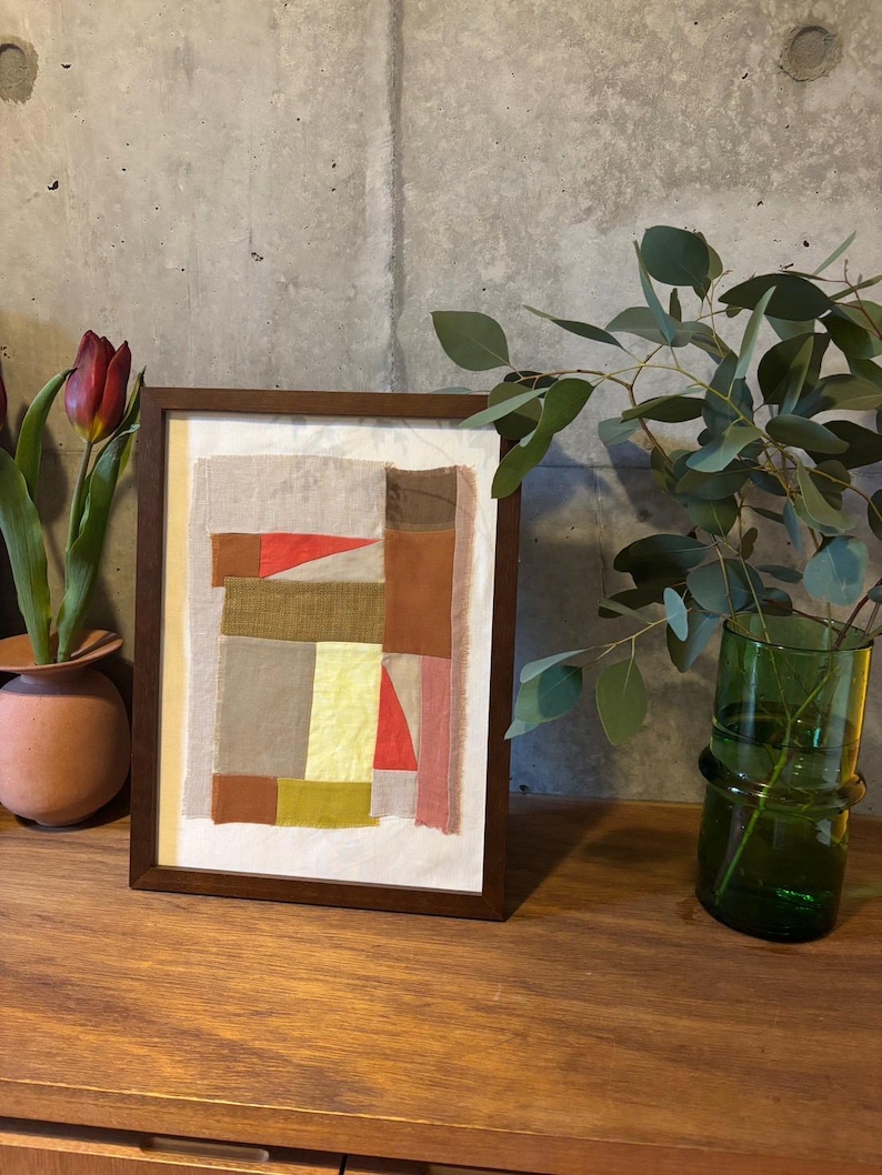 May include: A framed abstract art piece with a patchwork design of various shades of brown, beige, yellow, pink, and red fabric. The frame is dark brown wood.