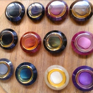 May include: Twelve decorative cabinet knobs arranged on a wooden surface. Each knob has a circular design with a central coloured insert, a gold-coloured ring, and a dark outer rim. Colours include black, red, purple, and white.