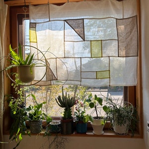 Puede incluir: Una ventana con una cortina de lino blanco con un patrón geométrico en tonos marrones y verdes. La ventana está decorada con varias plantas en macetas, incluyendo un cactus, un helecho y una planta de hojas.