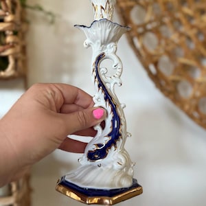 May include: White and gold ceramic candlestick holder with a blue and gold decorative accent. The candlestick holder has a detailed, ornate design with a flared base and a top designed to hold a candle. The holder is approximately 7 inches tall.