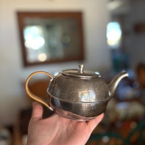 May include: A small, silver-colored teapot with a curved spout and a lid. The handle is a light tan color. The teapot is held in a person's hand.