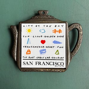 May include: A decorative metal teapot-shaped trivet with a white tile center. The tile features colorful illustrations and text: "CITY BY THE BAY, SAN FRANCISCO." Includes images of the sun, Golden Gate Bridge, and a cable car.